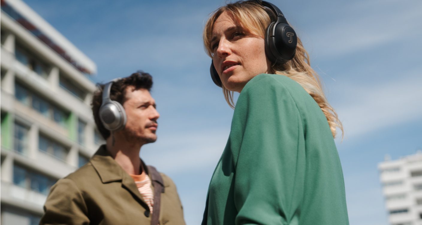 Two people listening to REAL BLUE PRO headphones in front of a blue sky and tall buildings.