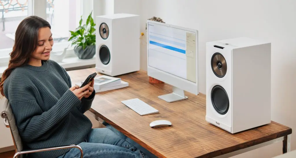 STEREO M2 Frau am Schreibtisch. Auf dem Schreibtisch steht ein Lautpsrecherpaar und ein Desktop. Sie schaut auf ihr Handy, um die Lautsprecher zusteuern.