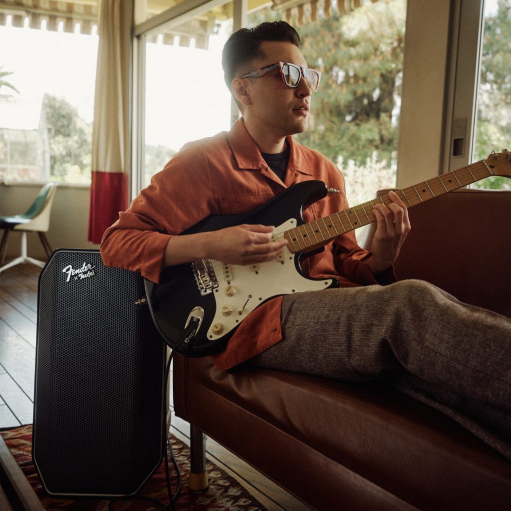 Mann sitzt auf Sofa mit E-Gitarre Fender Stratocaster in Schwarz, angeschlossen an Fender x Teufel ROCKSTER AIR 2